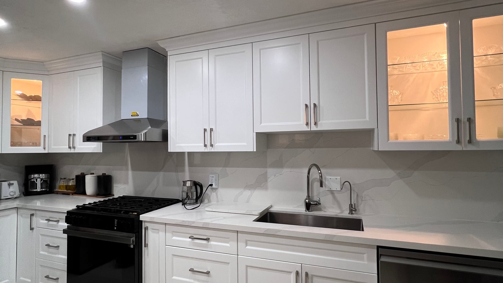 White kitchen cabinetry with illuminated glass-front upper doors, sink and appliance openings — manufactured in the Mississauga workshop (October 2026 catalogue)