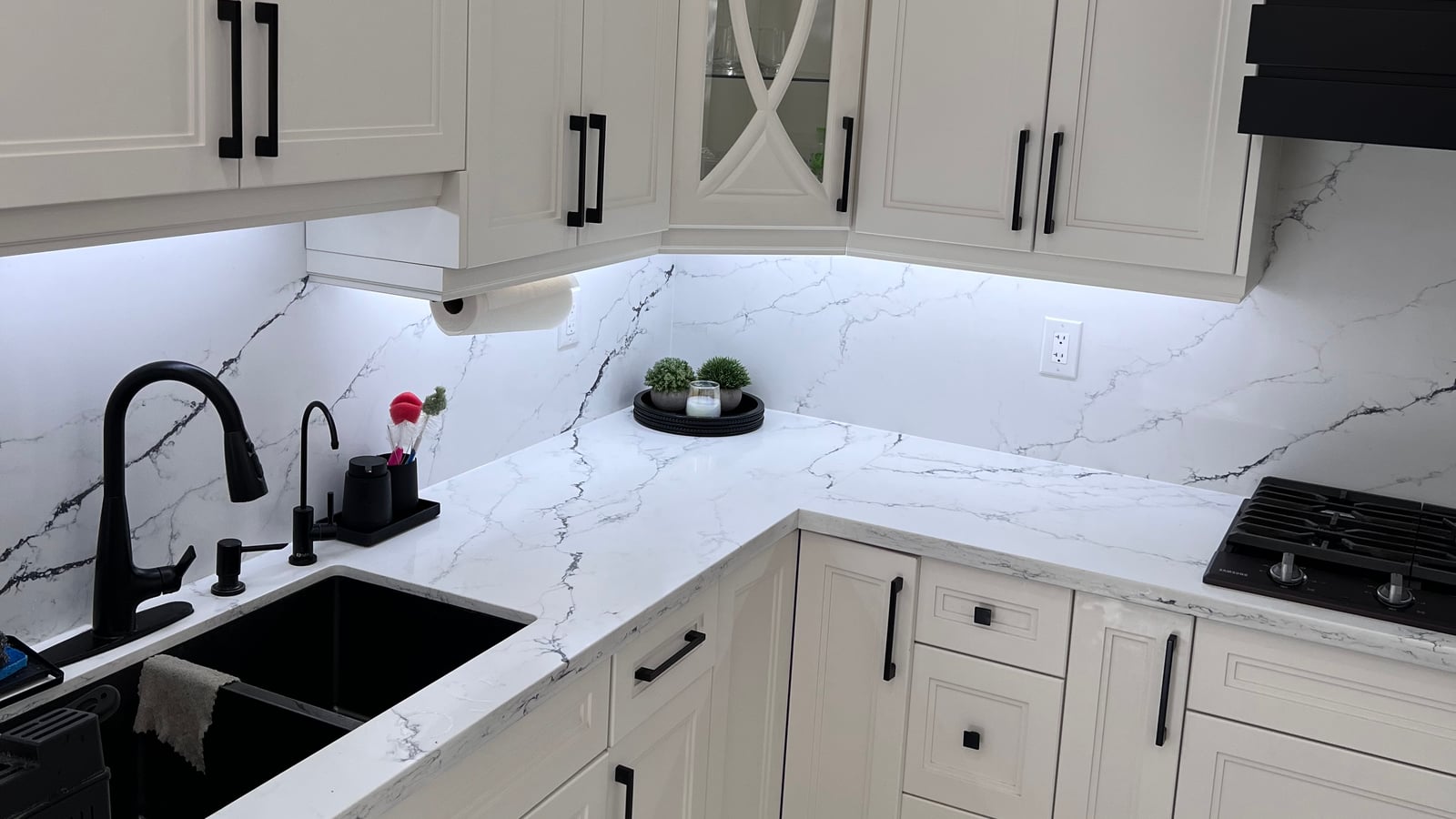 Kitchen with white cabinetry, glass-front display cabinets, a black decorative hood surround and veined countertop and backsplash surfaces — from Prime Kitchen and Bath's October 2026 catalogue