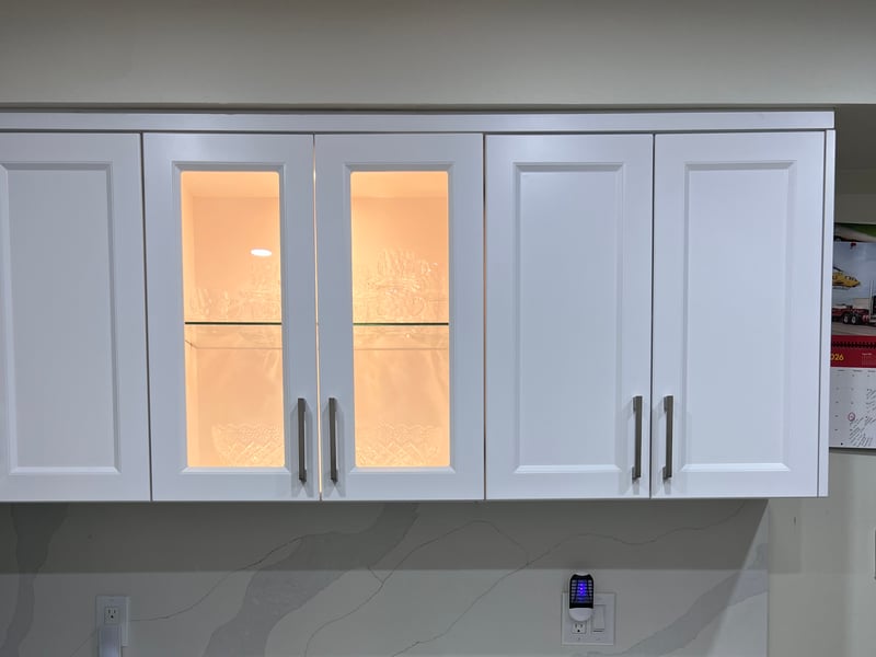 White kitchen cabinetry with illuminated glass-front upper doors, sink and appliance openings — manufactured in the Mississauga workshop (October 2026 catalogue)