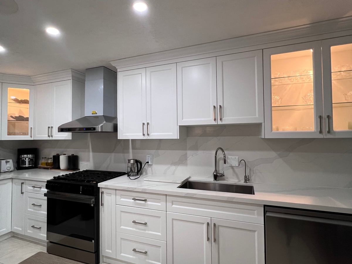 White kitchen with illuminated glass-front upper cabinets, a stainless hood and a marble-look quartz backsplash